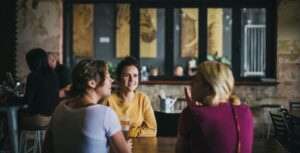 Three friends sharing a beer at a communal timber table inside Six Degrees Albany at dusk, mural and pendant lights visible behind.