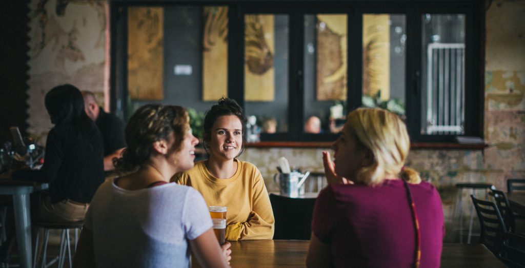 Three friends sharing a beer at a communal timber table inside Six Degrees Albany at dusk, mural and pendant lights visible behind.